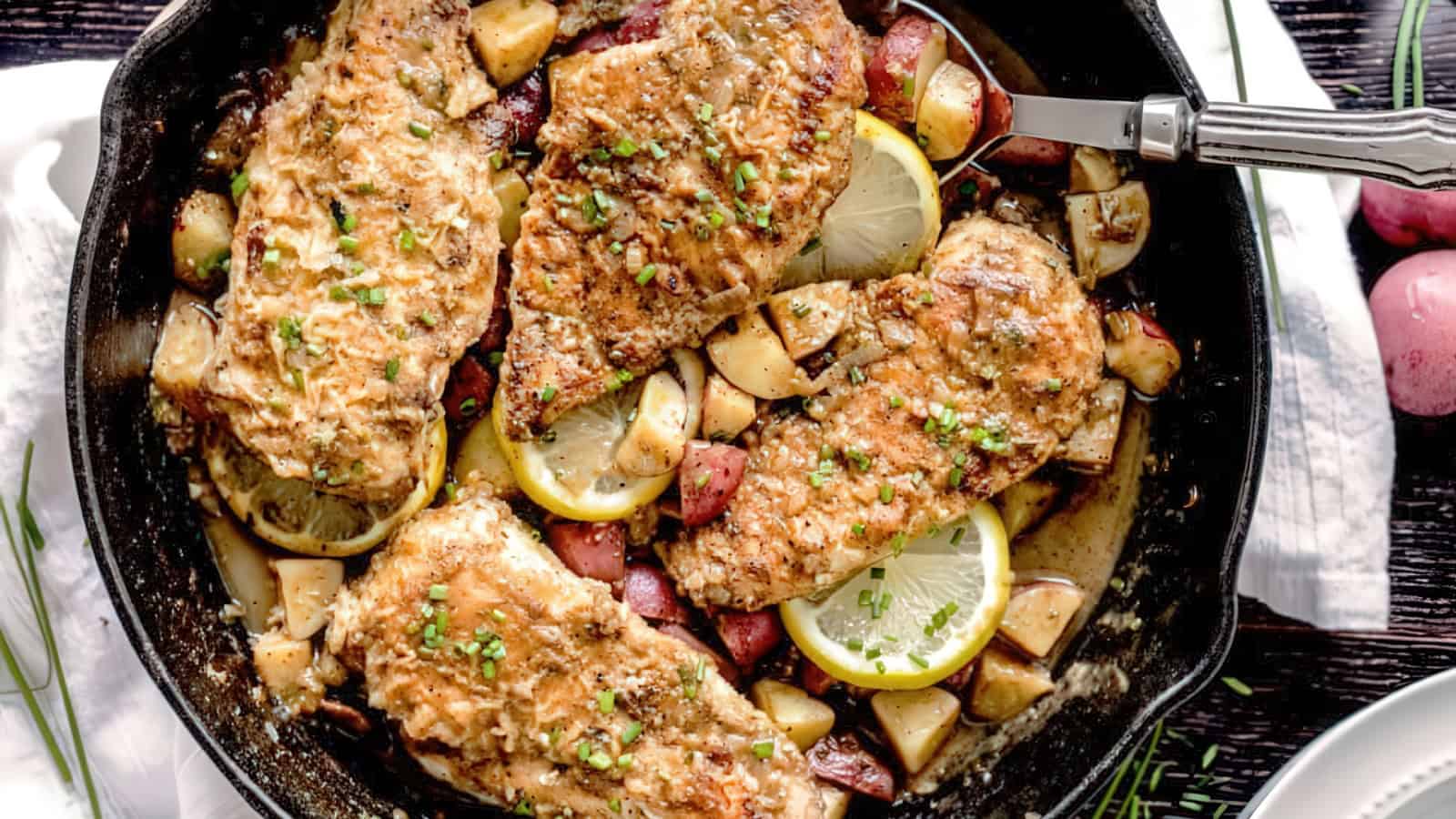 A skillet with four seasoned chicken breasts on top of diced potatoes. Lemon slices are tucked between the chicken, and the dish is garnished with chopped green herbs. A fork is placed in the skillet on the right side.
