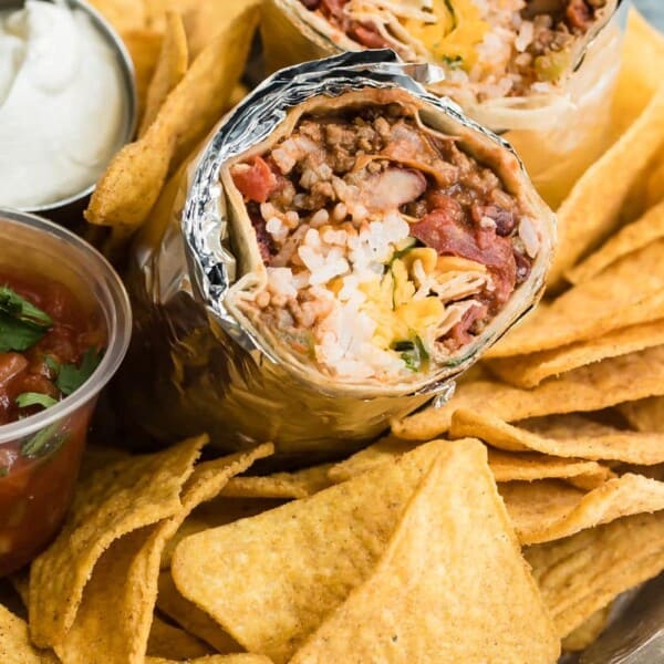 A tray of tortilla chips surrounds two foil-wrapped burritos filled with rice, beans, cheese, and ground meat. A small container of salsa garnished with cilantro and a dish of white sauce accompany the meal.