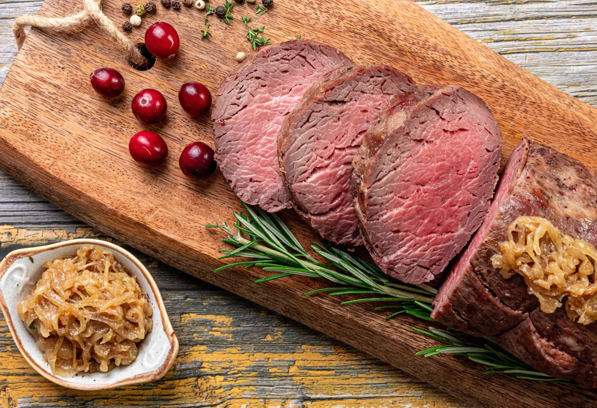 Sliced roast beef on a wooden board with rosemary sprigs, raw cranberries, peppercorns, and caramelized onions in a small dish.