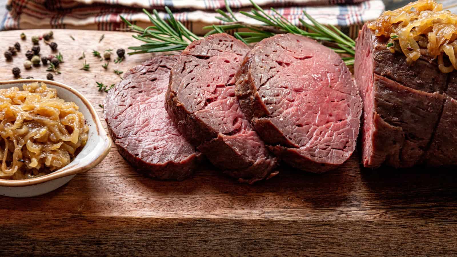 Sliced roast beef on a wooden cutting board, garnished with rosemary and served with caramelized onions on the side.