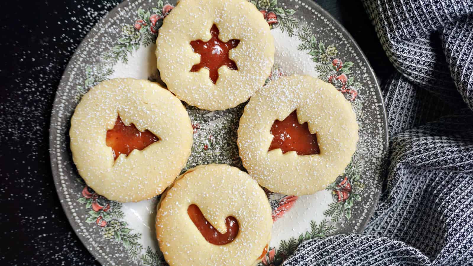 Four festive cookies with jam centers are arranged on a decorative plate. Each cookie has a unique cut-out: a star, Christmas tree, stocking, and candy cane. A gray patterned cloth drapes beside the plate.