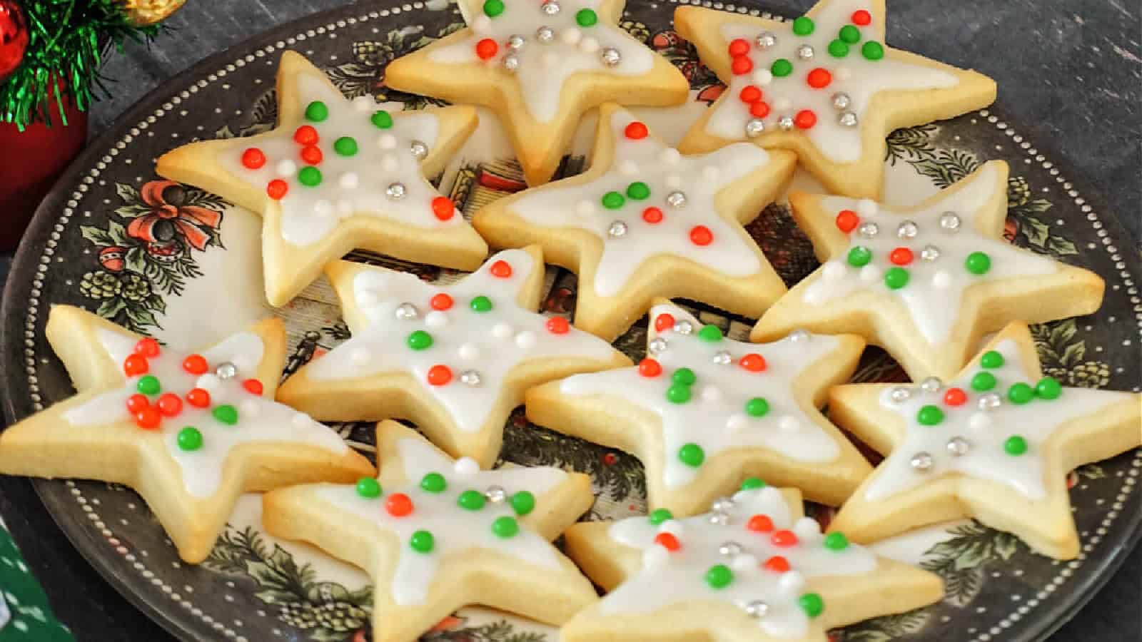 A plate of star-shaped cookies decorated with white icing and topped with red, green, and silver sprinkles. The plate has a festive design and is set on a dark surface, suggesting a holiday theme.