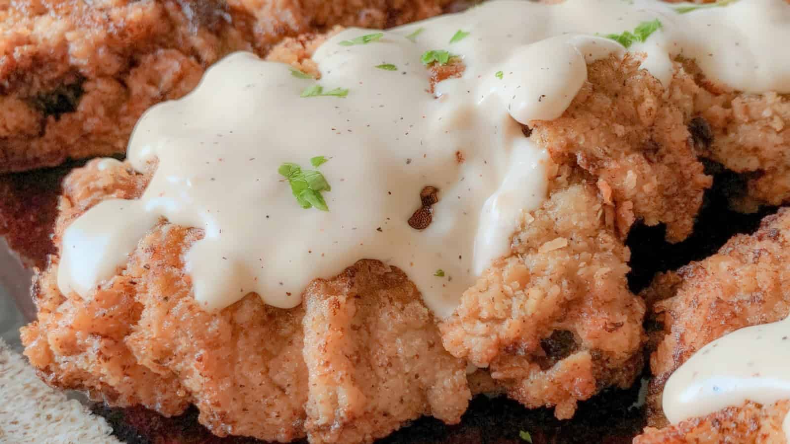 Close-up of crispy fried chicken smothered in creamy white gravy, garnished with chopped herbs. The golden, crunchy texture of the chicken contrasts with the smooth, rich sauce.