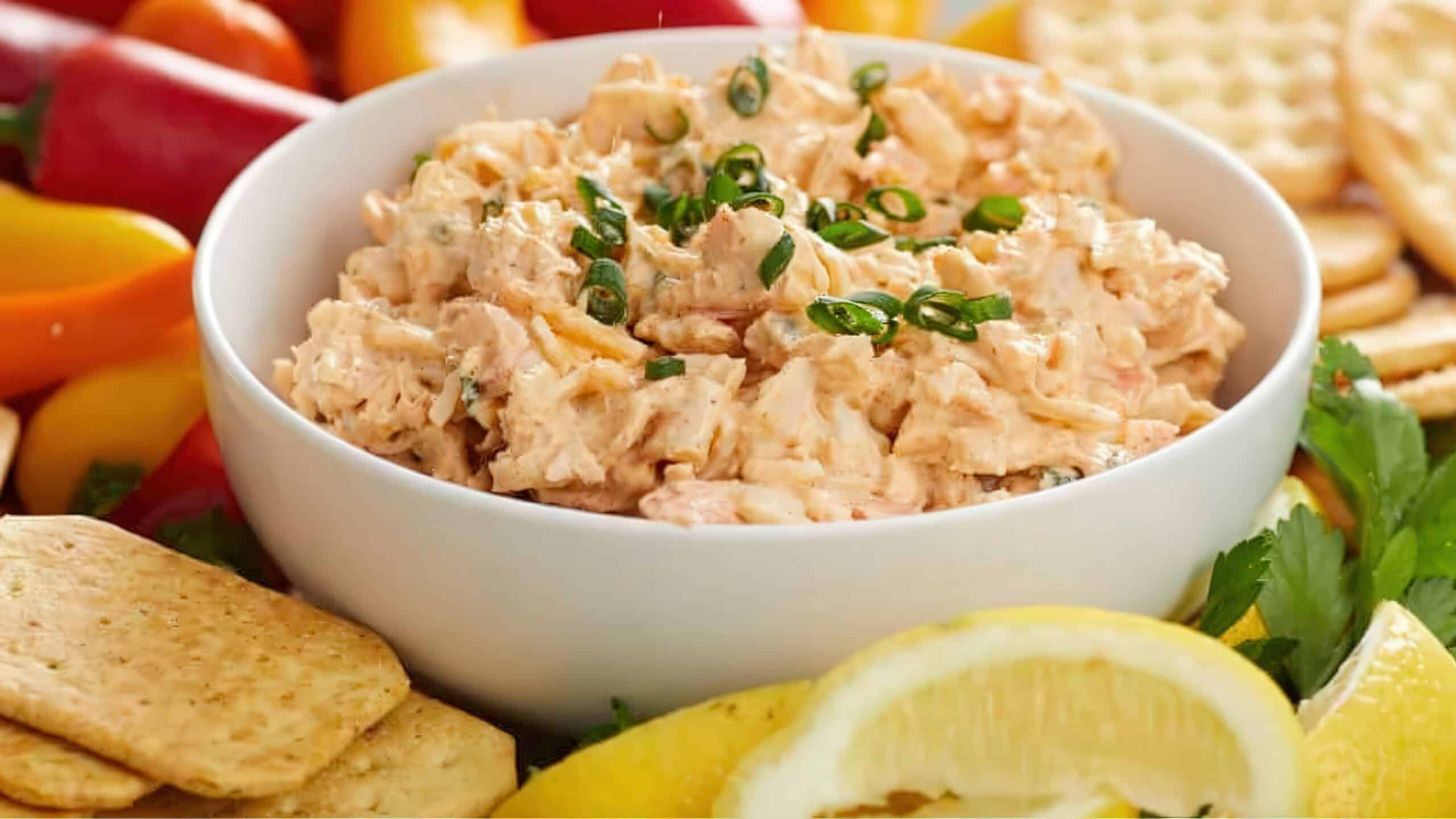 A white bowl filled with creamy salmon dip, garnished with chopped green onions, surrounded by crackers, sliced lemons, and colorful peppers on a platter.