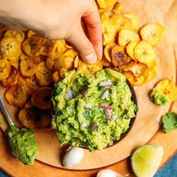 Hand dipping a plantain chip into guacamole, with more chips, cilantro leaves, a garlic clove, and lime wedge on a wooden board.