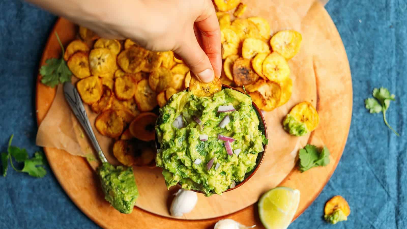 A hand dips a plantain chip into a bowl of guacamole, surrounded by a plate of fried plantains. Lime, garlic, and cilantro garnish the blue tablecloth backdrop.