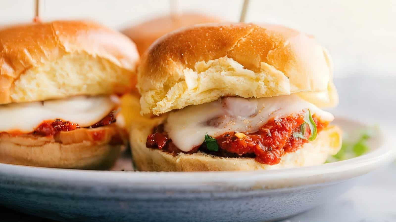 Close-up of a chicken parmesan slider on a white plate. The sandwich features a breaded chicken cutlet topped with marinara sauce and melted cheese, encased in a soft golden bun. Another slider is partially visible in the background.