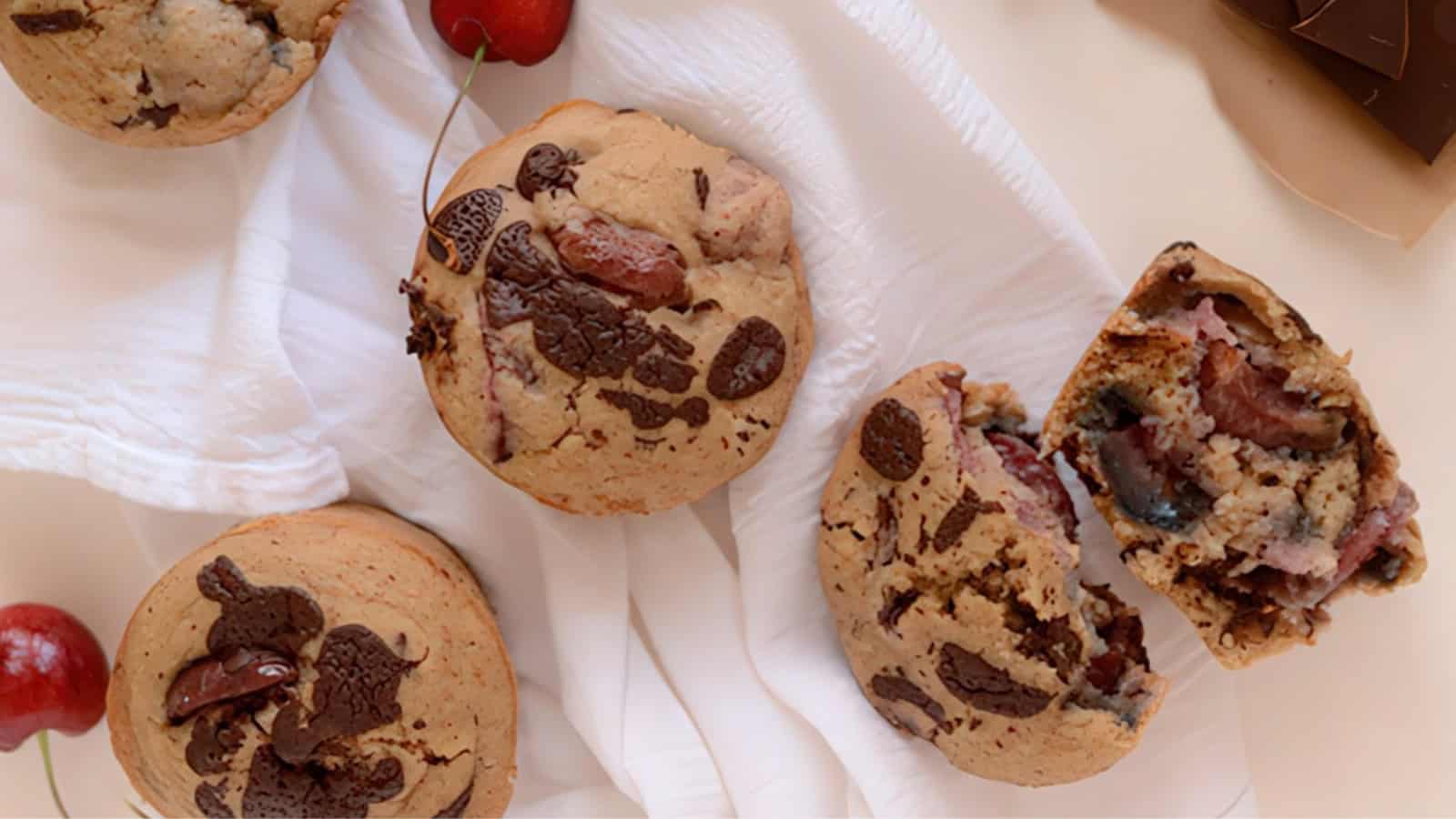 Top view of freshly baked muffins with chocolate chunks on a white cloth. One muffin is cut in half, revealing a moist interior with visible fruits and chocolate. A few cherries and a piece of chocolate are scattered around the scene.