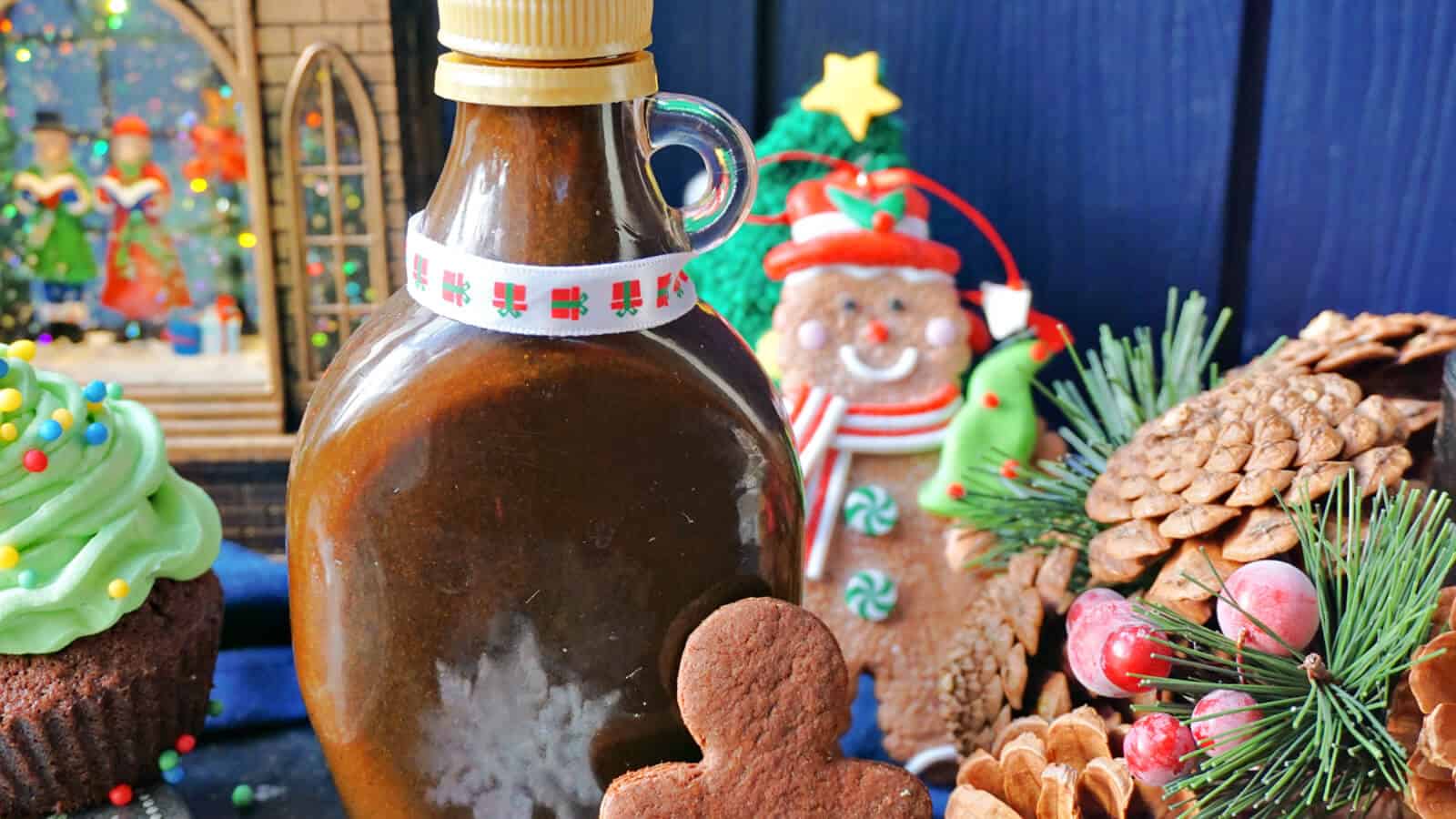 A festive holiday scene featuring a bottle of syrup, Christmas cookies, a green cupcake, a gingerbread man, pinecones, and a small Christmas tree. A decorative holiday backdrop completes the cozy, seasonal arrangement.