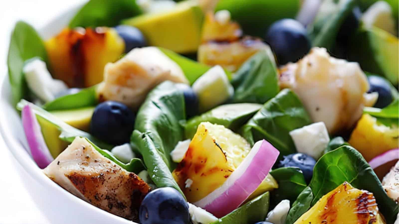 A fresh salad in a white bowl featuring grilled chicken, pineapple chunks, blueberries, avocado, red onion slices, and spinach leaves.