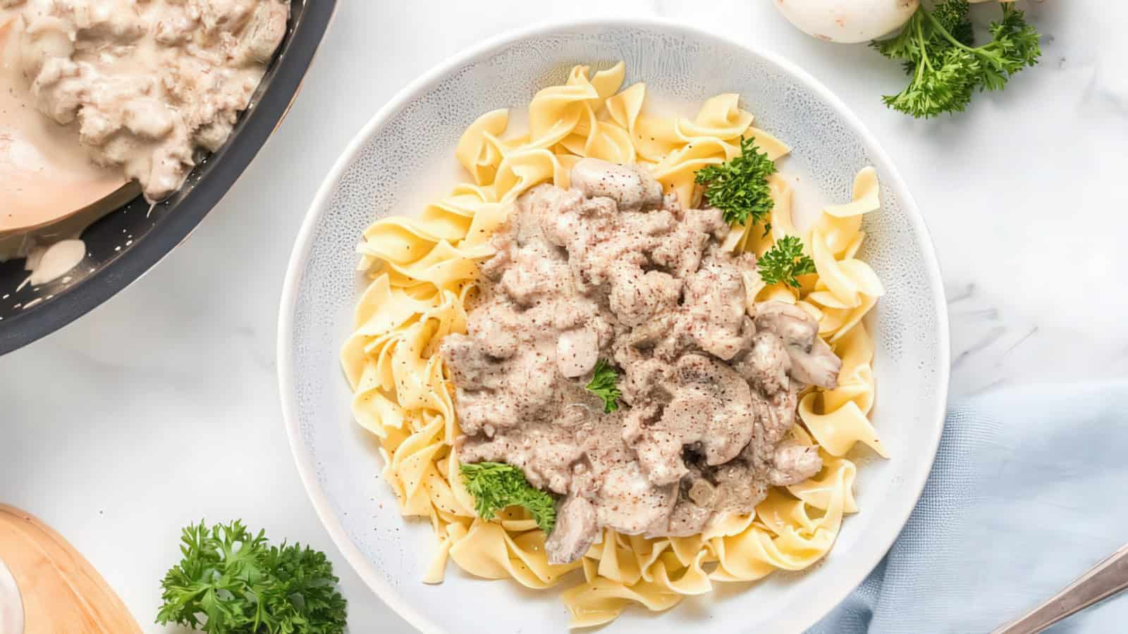 A plate of creamy beef stroganoff on top of egg noodles, garnished with fresh parsley. Nearby, a pan with more stroganoff and parsley sprigs rest on a marble countertop.