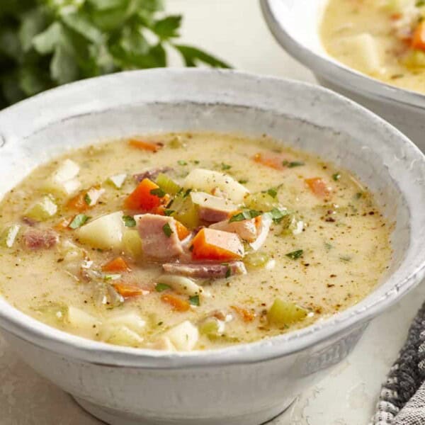 A bowl of creamy soup with diced ham, potatoes, carrots, and herbs on a table beside a gray cloth and fresh parsley.