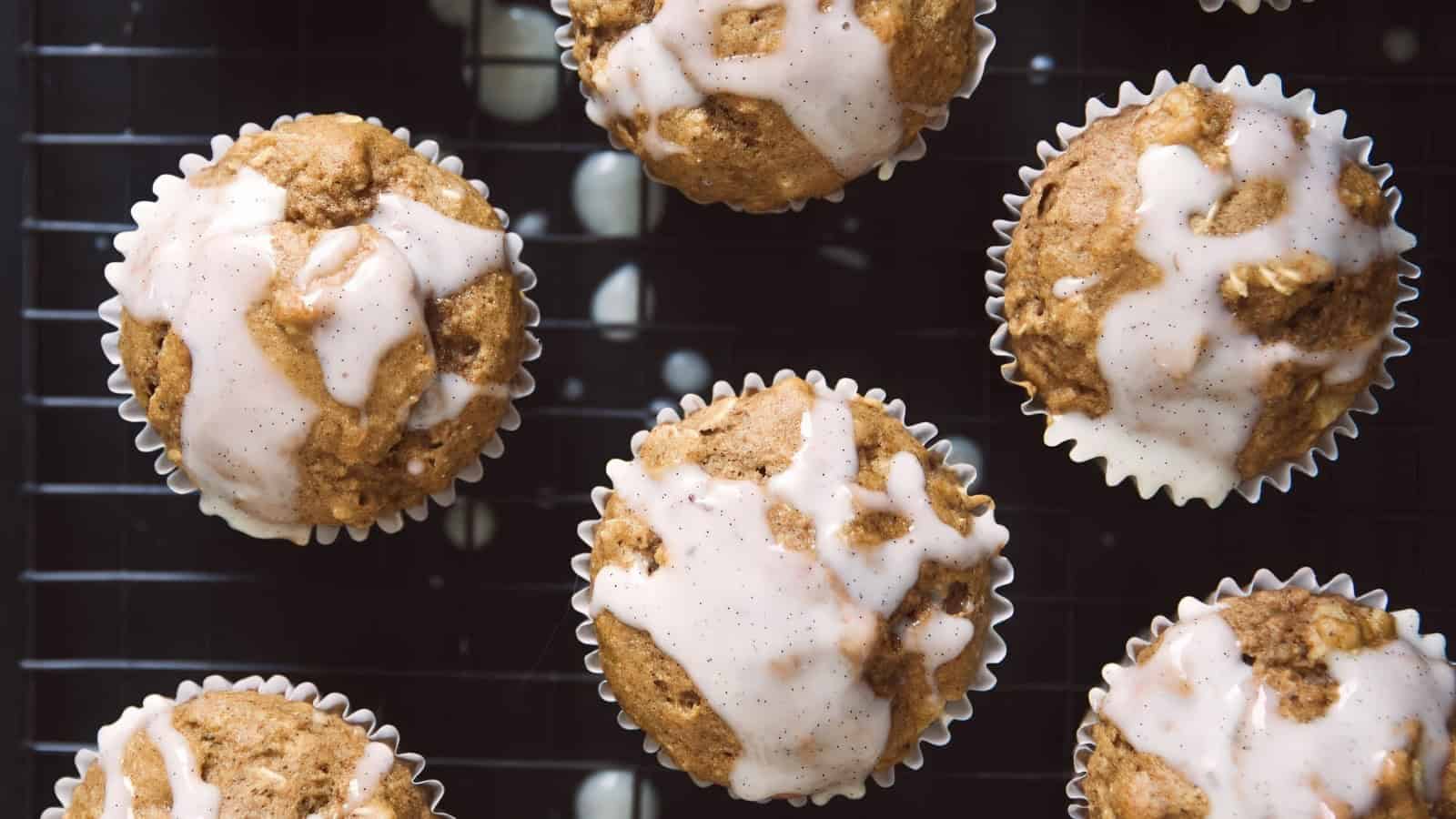 Seven muffins with a light glaze on top are arranged on a cooling rack. The muffins are golden-brown and have a textured, crumbly appearance. The glaze is drizzled unevenly, adding a sweet contrast to their rustic look.
