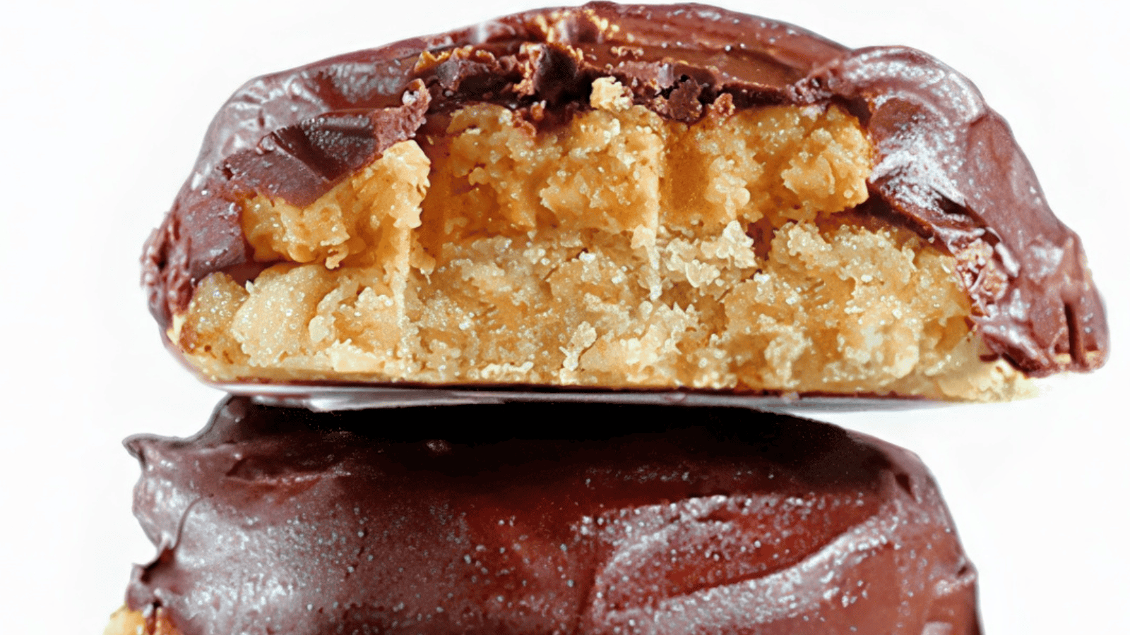 Close-up of a halved chocolate-covered peanut butter cookie with a crumbly texture. The top piece shows a smooth chocolate coating and a soft, golden peanut butter filling, providing a rich, indulgent look against a white background.