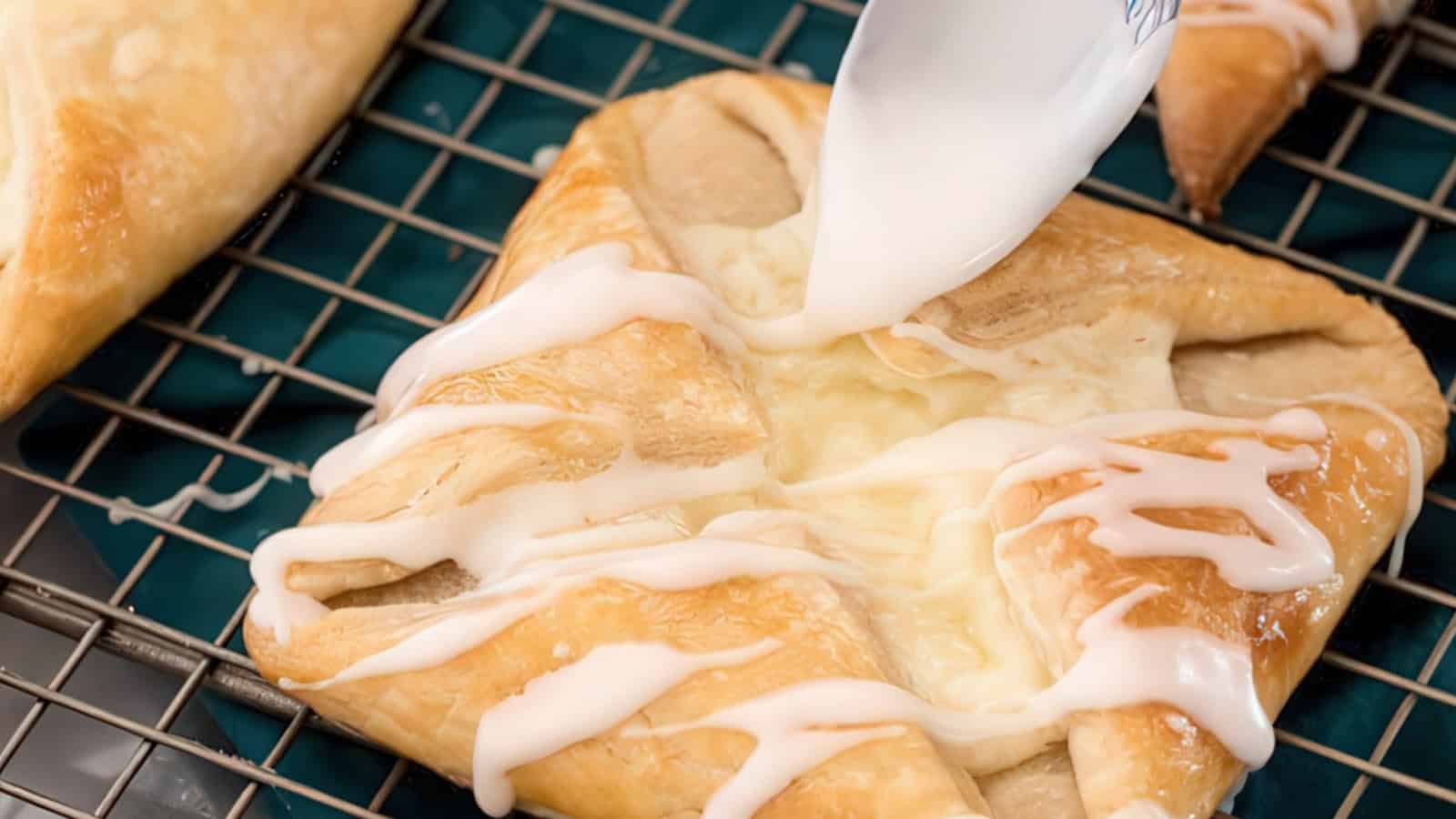 A close-up of a pastry with a golden, flaky crust being drizzled with white icing. The pastry rests on a cooling rack over a blue surface.