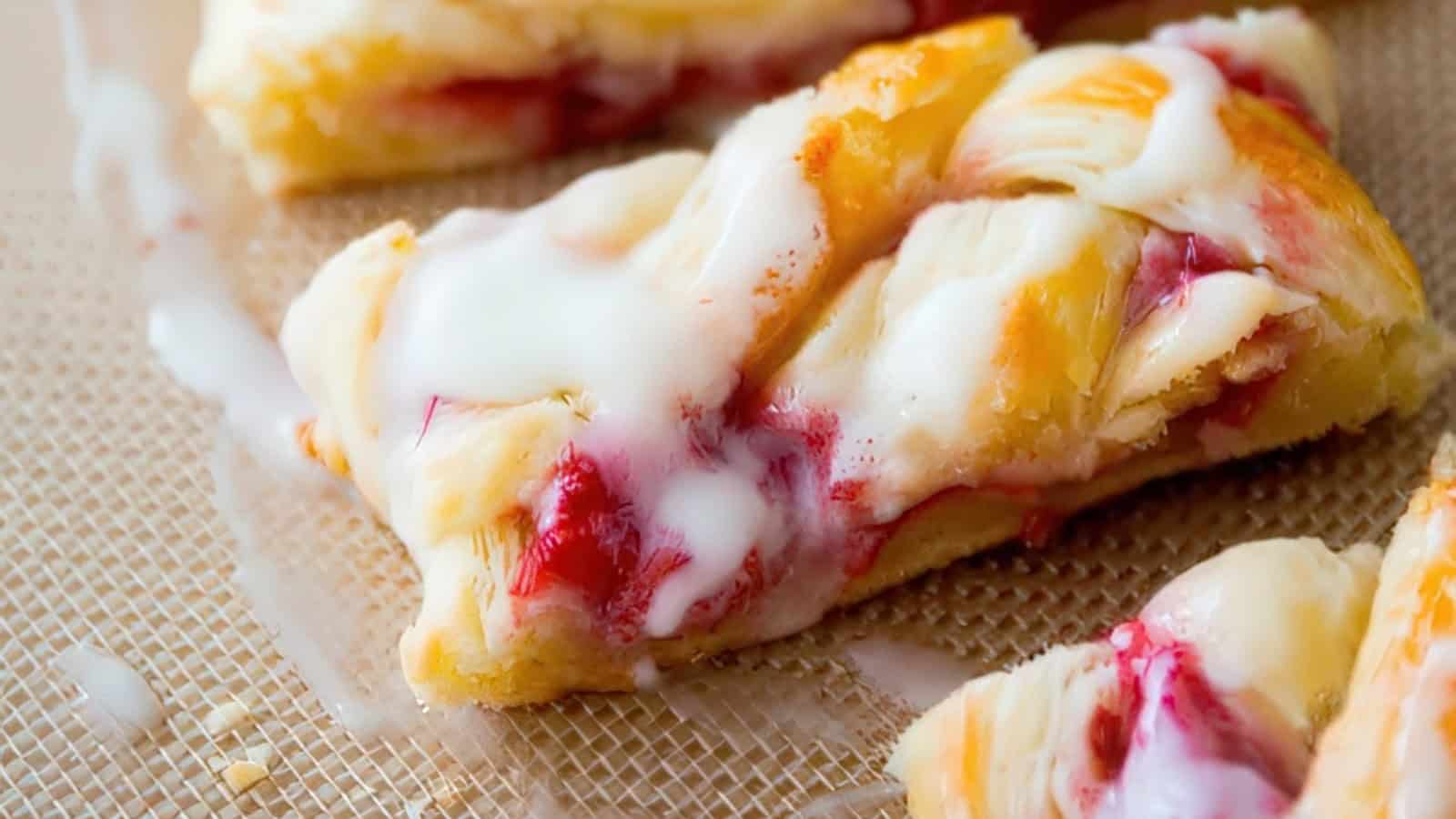 A close-up of a pastry with a braided texture and a drizzle of white icing. The pastry has a golden-brown crust and a filling of red berry jam, placed on a textured baking mat.