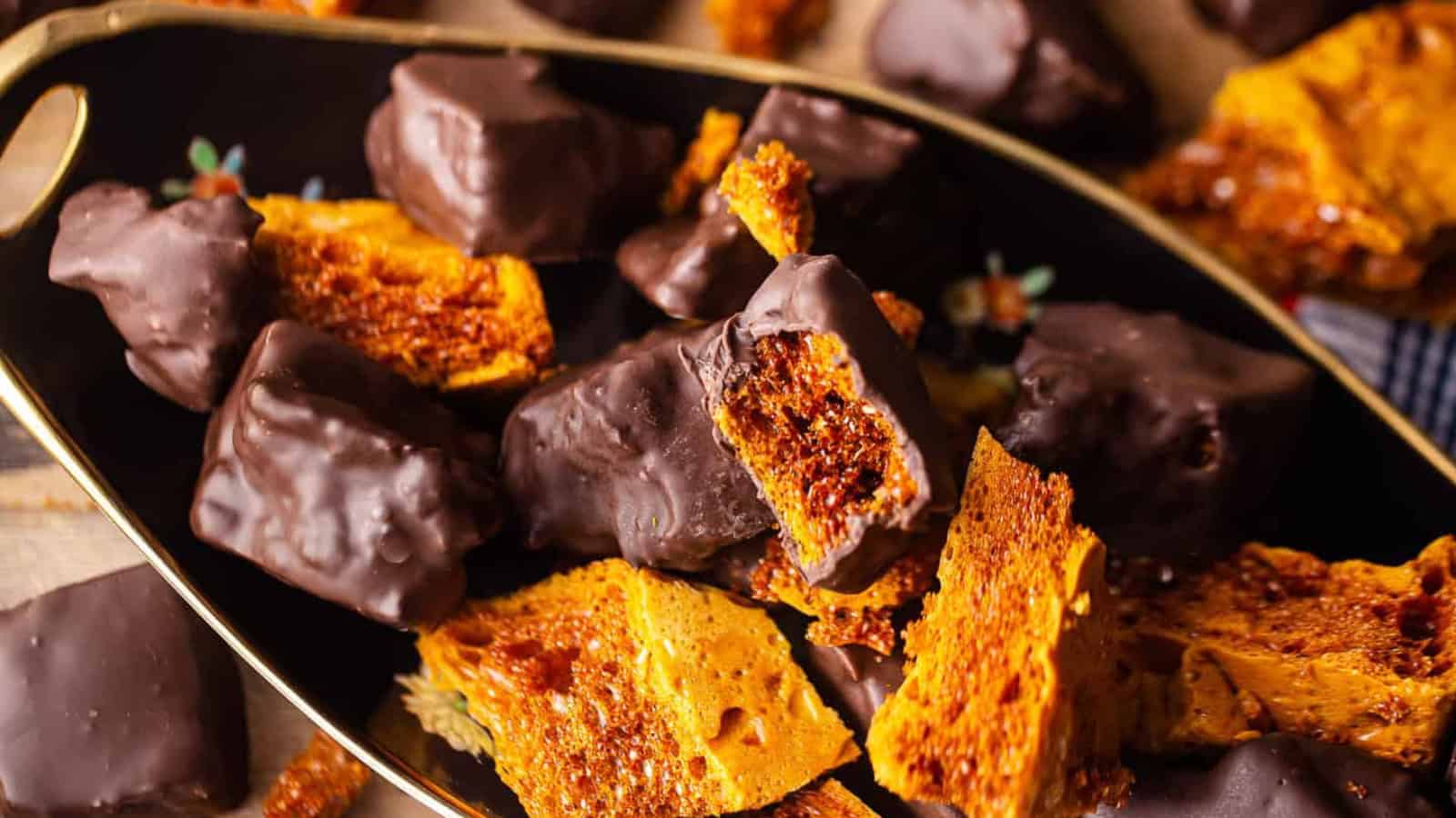 A black dish filled with chocolate-covered honeycomb pieces mixed with plain honeycomb chunks. The golden honeycomb has a crispy texture, while the chocolate coating is smooth and glossy.