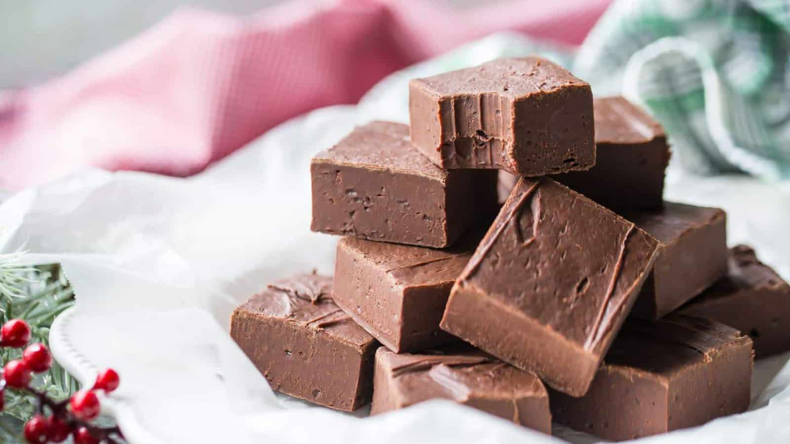 A stack of chocolate fudge squares arranged on a white parchment paper. The background includes hints of a red and green cloth, and a sprig of holly with red berries, suggesting a festive setting.