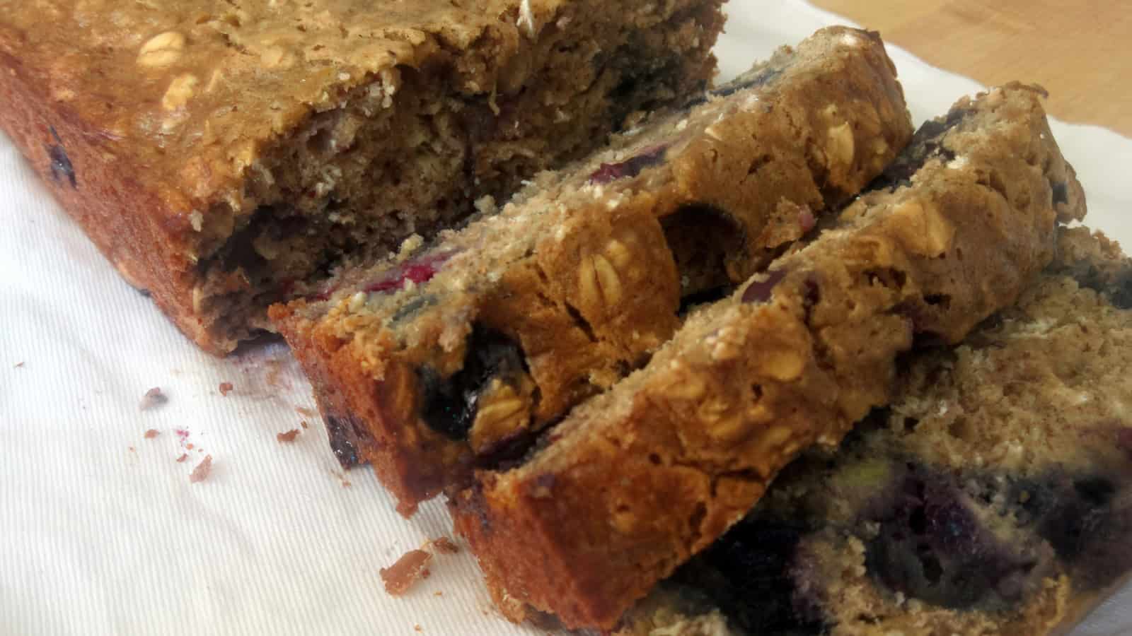 A loaf of sliced banana bread with visible blueberries and nuts, placed on a white napkin. The bread appears moist and dense, showcasing a golden-brown crust and a soft interior.