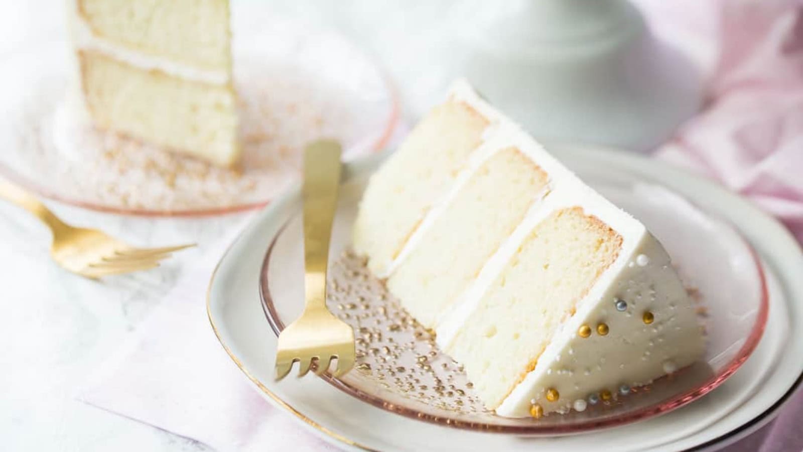 A slice of three-layer vanilla cake with white frosting on a plate, garnished with gold and silver sprinkles. A gold fork rests beside the plate. There is another slice of cake in the background. The setting is soft and elegant.
