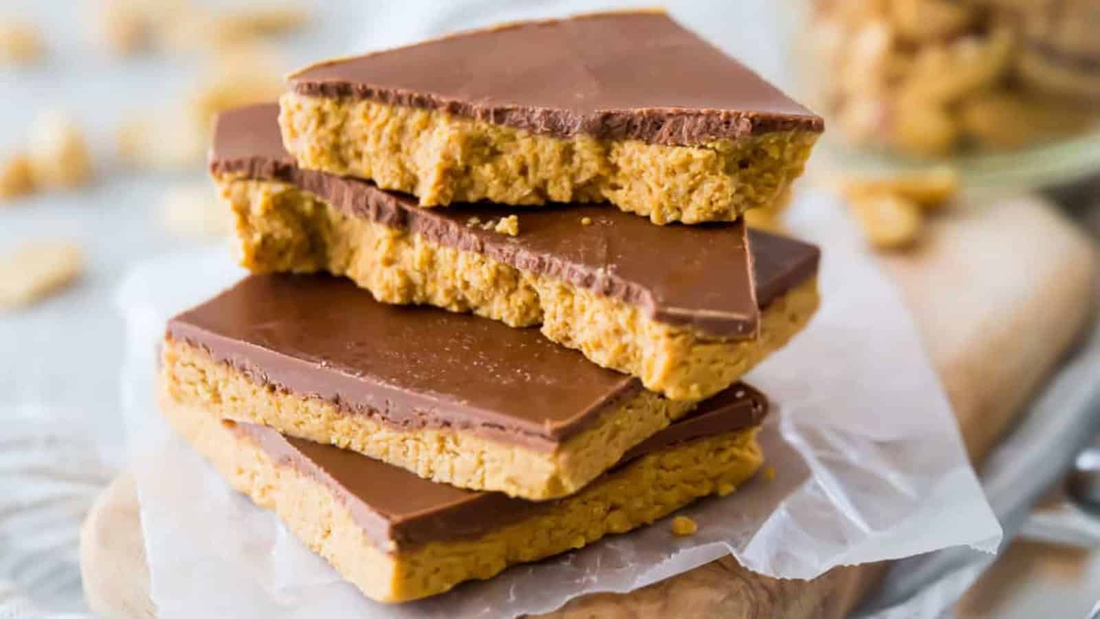 Four triangular peanut butter bars with a chocolate topping are stacked on a piece of parchment paper. The bars are placed on a wooden cutting board, with more peanut butter pieces blurred in the background.