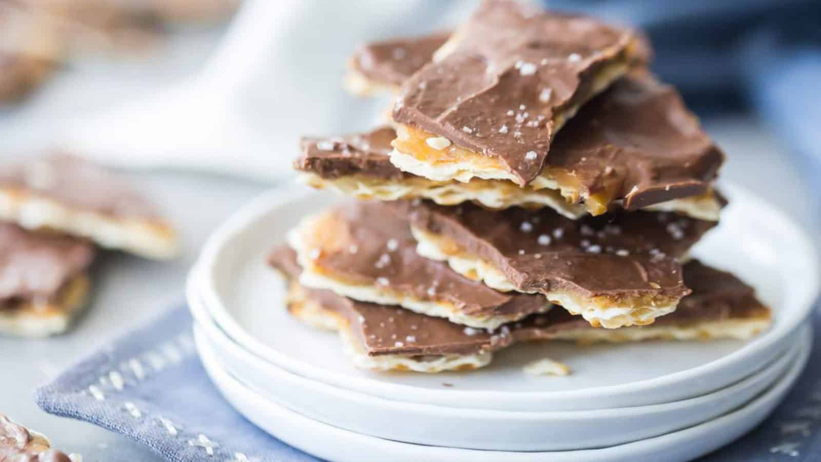 A stack of chocolate-covered toffee pieces rests on a white plate. The toffee has a golden crust and is topped with a layer of rich chocolate, sprinkled with sea salt. The background is softly blurred with a hint of a blue fabric.