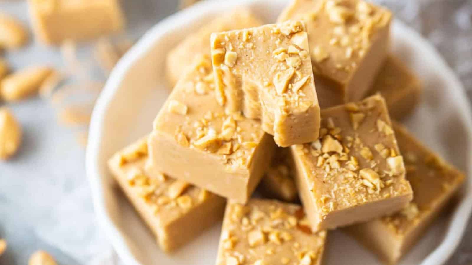 A stack of creamy peanut butter fudge squares topped with chopped peanuts, arranged on a white plate. The background is softly blurred, showcasing more peanuts scattered around.