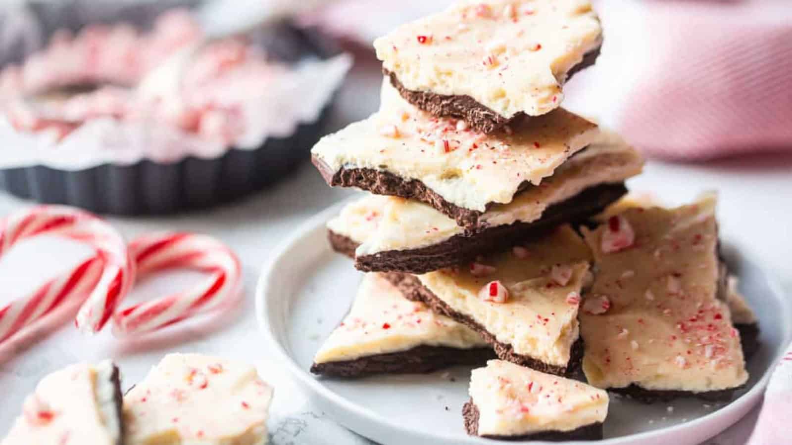 A stack of peppermint bark pieces on a white plate. The bark features layers of dark chocolate and white chocolate, sprinkled with crushed peppermint candies. Two candy canes and more bark are in the background.