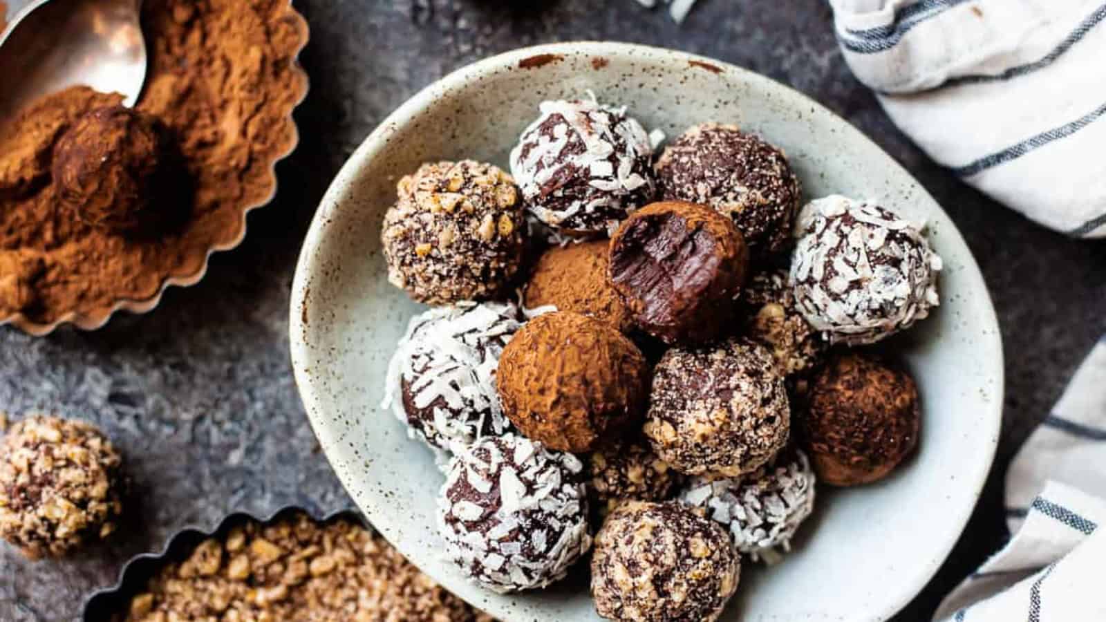 A bowl of assorted chocolate truffles coated with coconut, cocoa powder, and crunchy bits. Some truffles are placed in small dishes nearby, with scattered ingredients, all set on a dark textured surface.