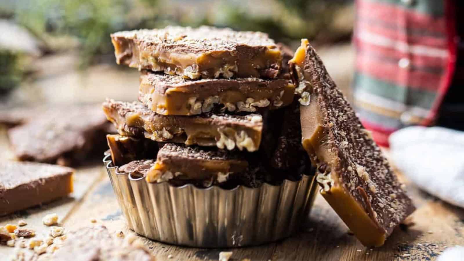 A pile of homemade toffee pieces with chocolate and nuts in a small metal dish, set on a rustic wooden surface. A checkered cloth is visible in the background, adding a cozy touch.