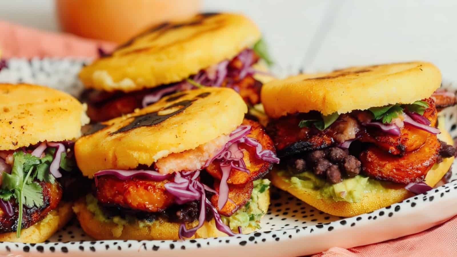 Three arepas stuffed with roasted plantains, black beans, avocado, shredded red cabbage, and cilantro on a white plate with black spots. A soft orange napkin is partially visible underneath the plate.