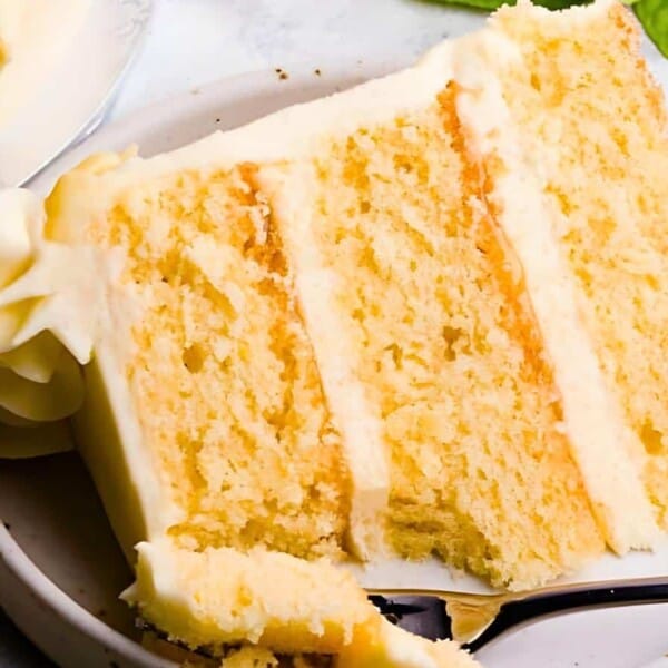 A slice of three-layer yellow cake with white frosting on a plate and a fork holding a piece.