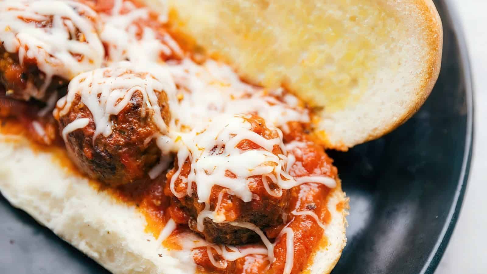 A meatball sub on a black plate features several meatballs covered in marinara sauce and melted cheese, nestled in a toasted sub roll.