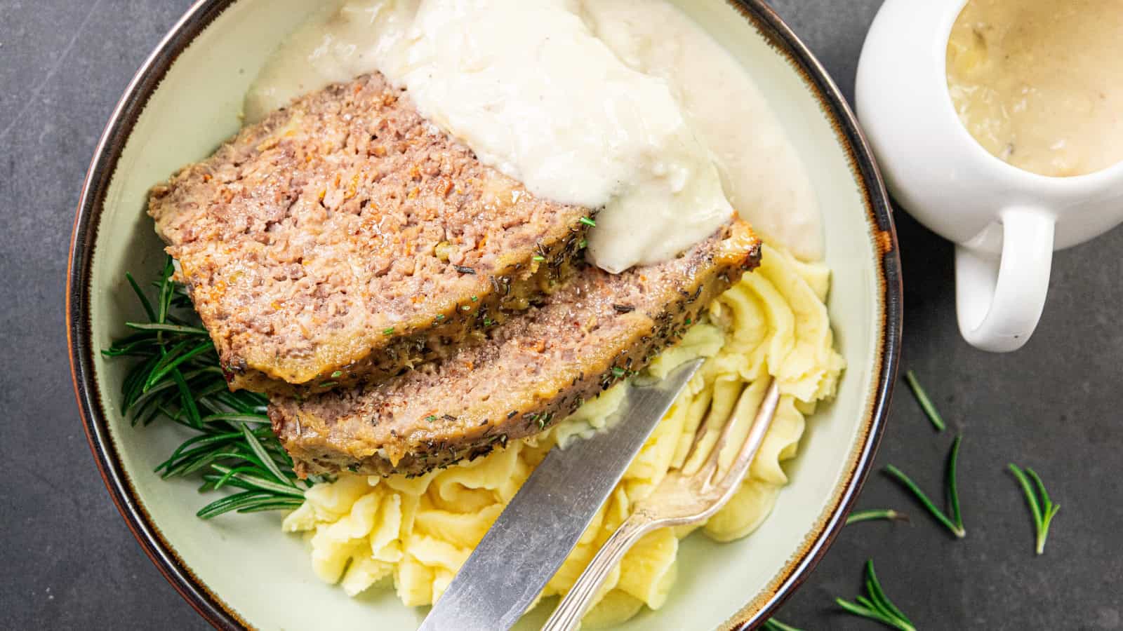 A plate with two slices of meatloaf topped with gravy, served on a bed of mashed potatoes. A sprig of rosemary garnishes the dish, with a fork and knife alongside. A white gravy boat is partially visible on the side.