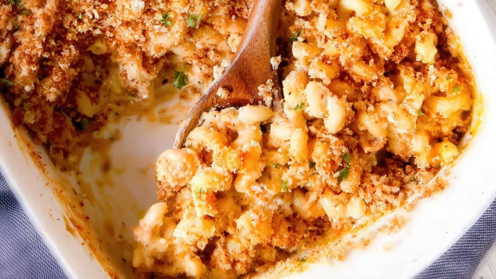 A close-up of a baking dish containing creamy macaroni and cheese, topped with a crispy golden breadcrumb crust. A wooden spoon is scooping out a portion, revealing the cheesy interior.