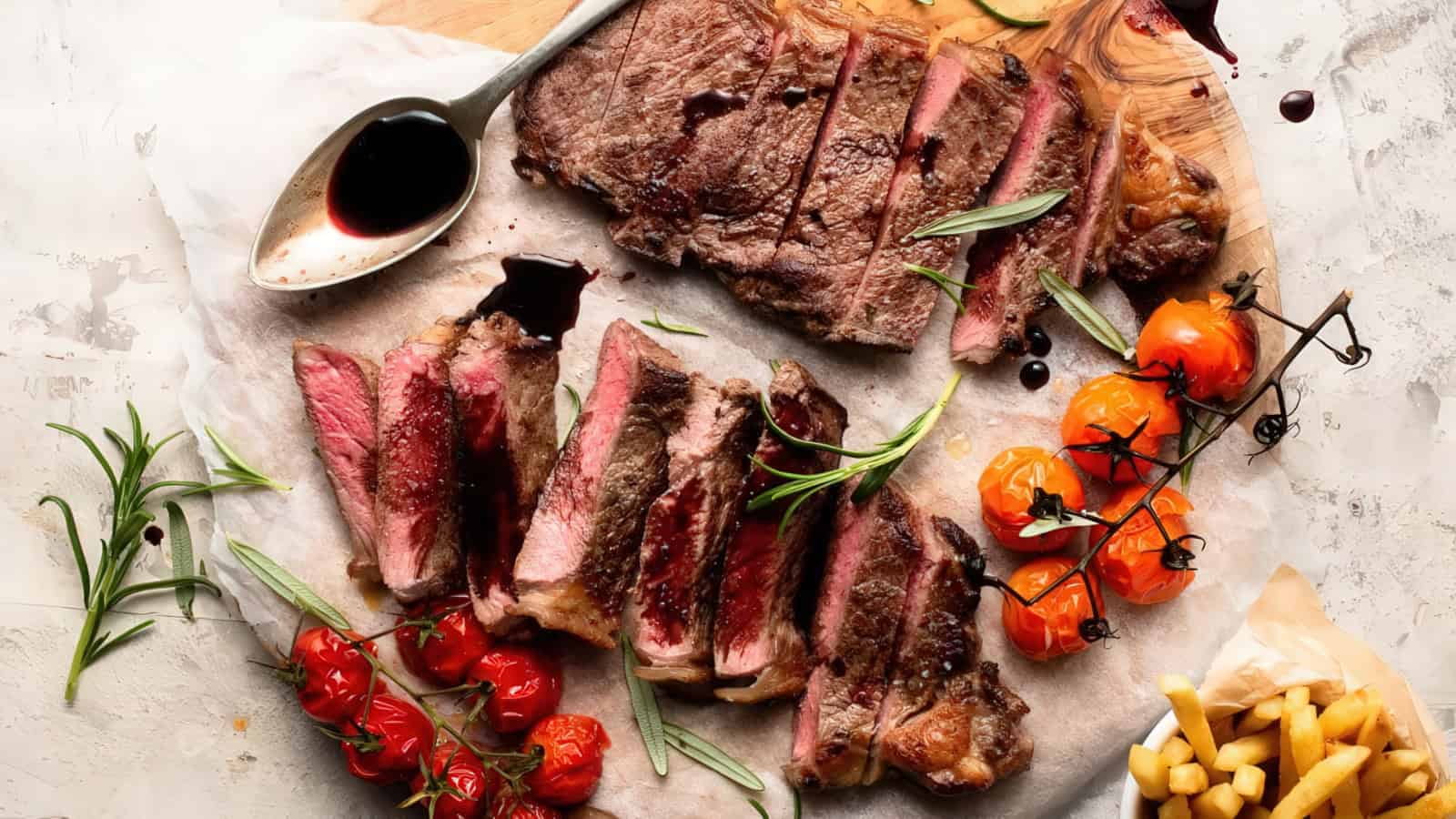 A sliced, medium-rare steak with a dark sauce drizzled on top, surrounded by sprigs of rosemary. Cherry tomatoes on the vine and a serving of golden French fries are arranged alongside on a wooden board.