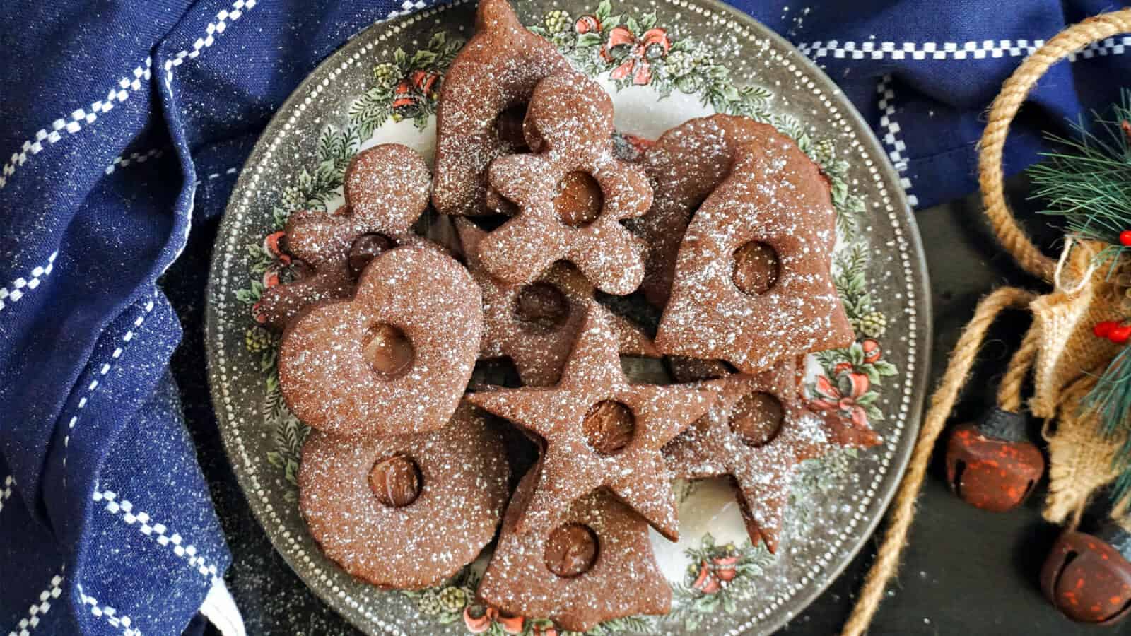 A plate of assorted chocolate cookies in various shapes, including stars, hearts, and flowers, dusted with powdered sugar. The plate has a floral design and rests on a dark surface near a blue cloth with white stitching.