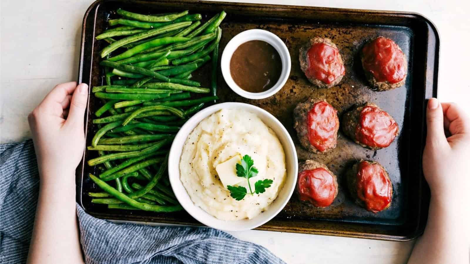 A tray with roasted green beans, mashed potatoes topped with butter and parsley, a bowl of brown gravy, and six mini meatloaves with ketchup glaze. Hands are holding the tray on a striped cloth.