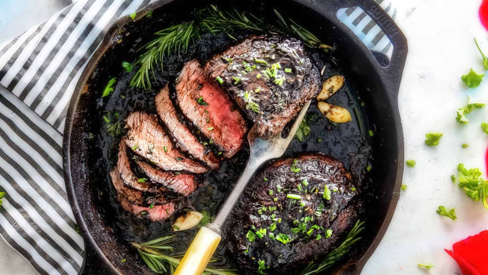 A cast iron skillet with sliced medium rare steak garnished with herbs and garlic cloves. Fresh rosemary sprigs and chopped parsley are sprinkled on top. A striped cloth and parsley leaves are visible beside the skillet.
