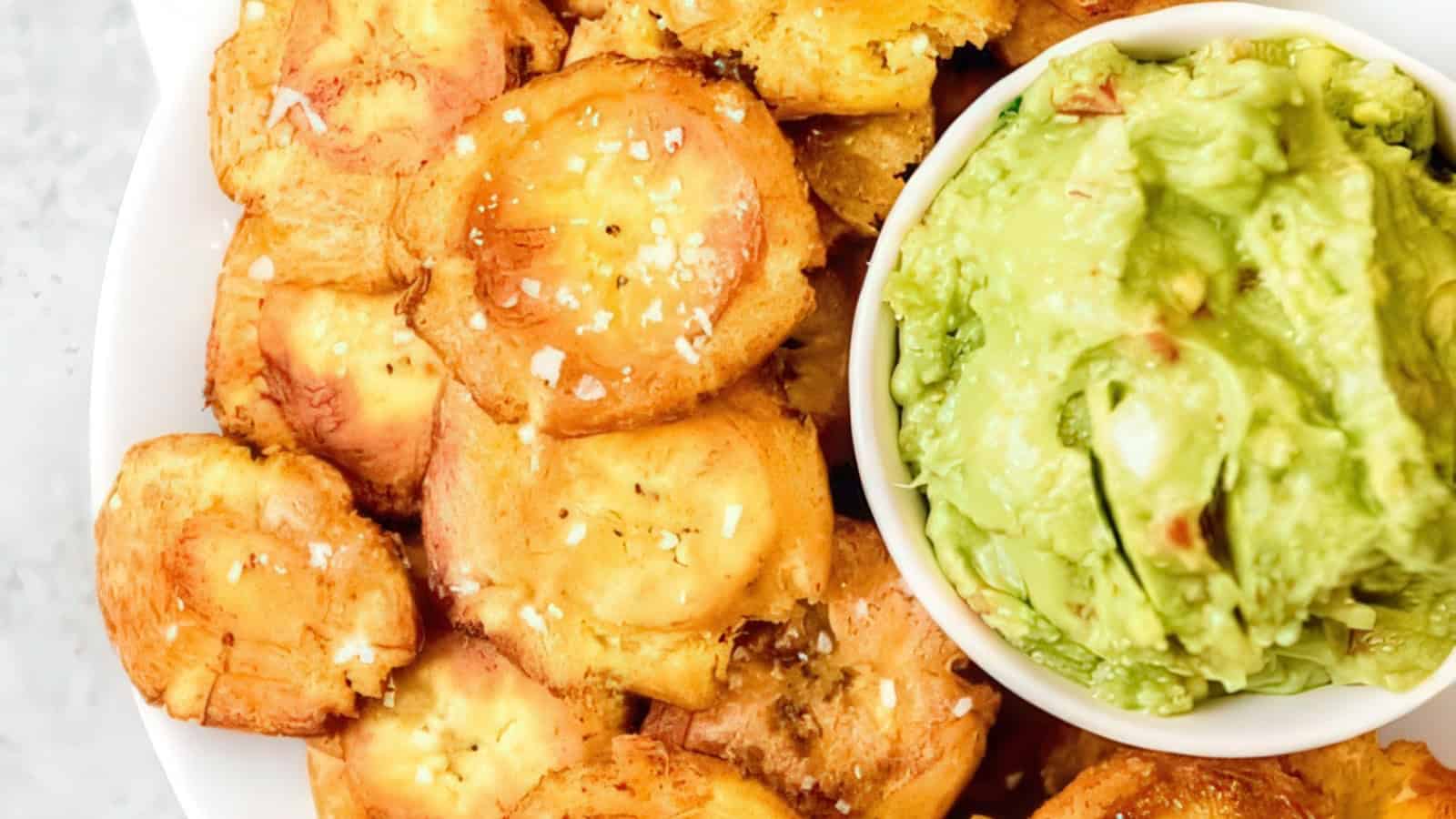 A plate of golden, crispy tostones sprinkled with salt is shown next to a bowl of creamy guacamole.