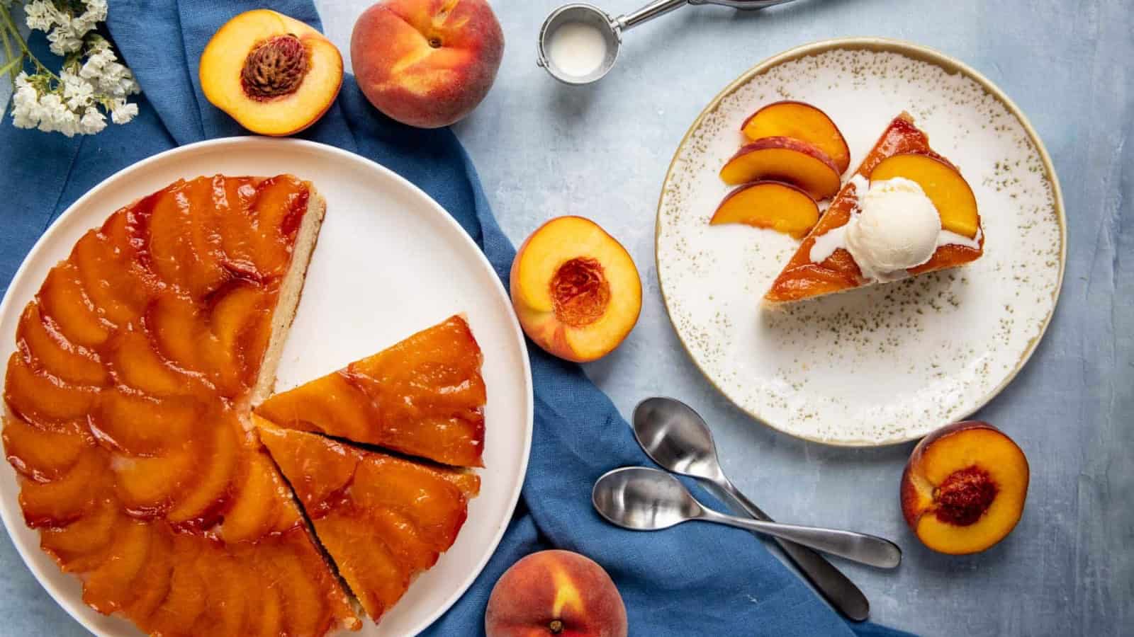A glazed peach upside-down cake on a white plate with a slice served on a smaller plate. The slice is topped with a scoop of ice cream. Fresh peaches and two spoons are nearby on a blue cloth.
