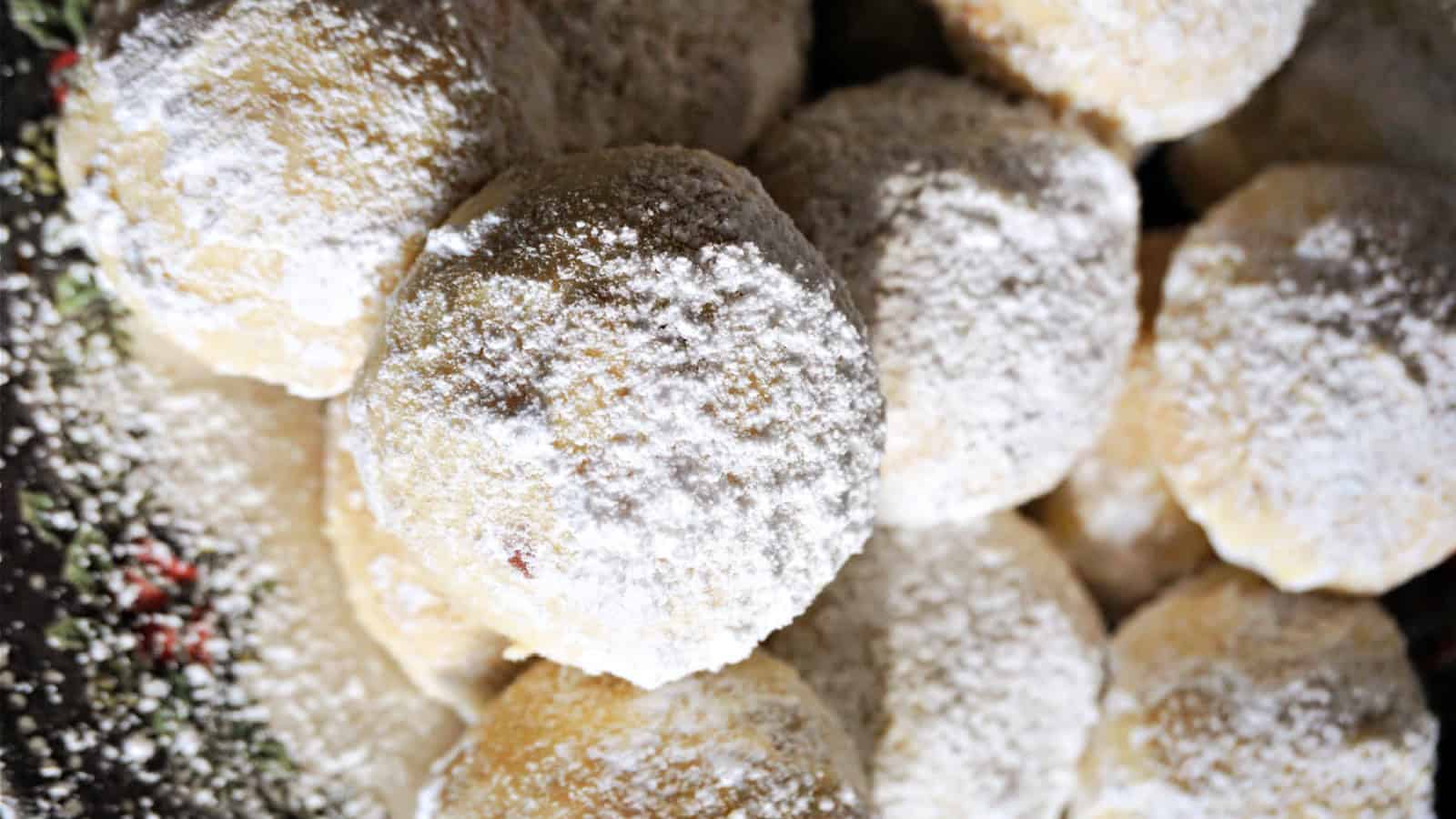 A close-up of several round cookies covered in powdered sugar, with a slightly rough texture. The cookies are piled on top of each other, creating a festive and appetizing display.