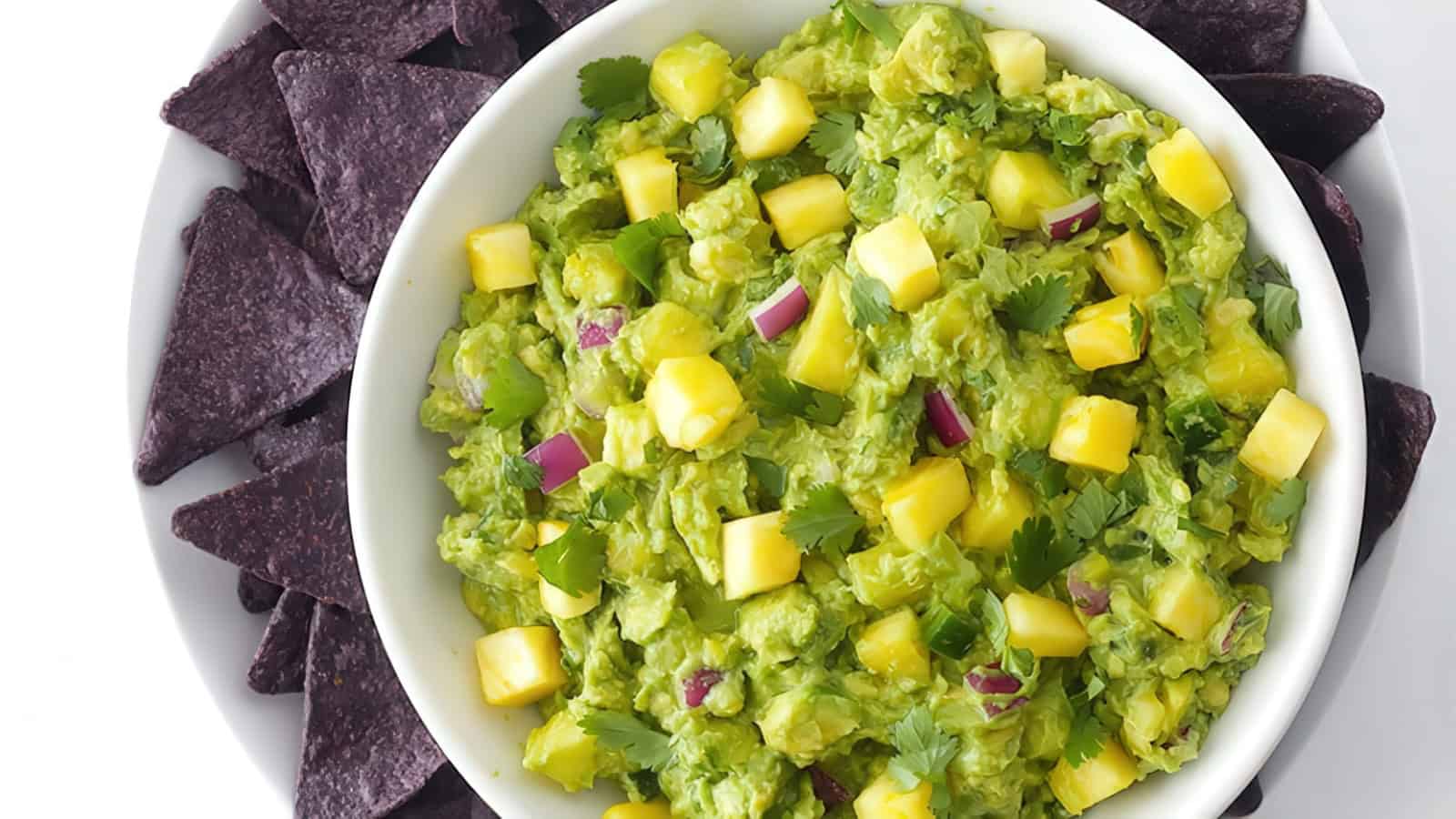 A bowl of guacamole with mango chunks, red onion, and cilantro, surrounded by blue corn tortilla chips on a white plate.