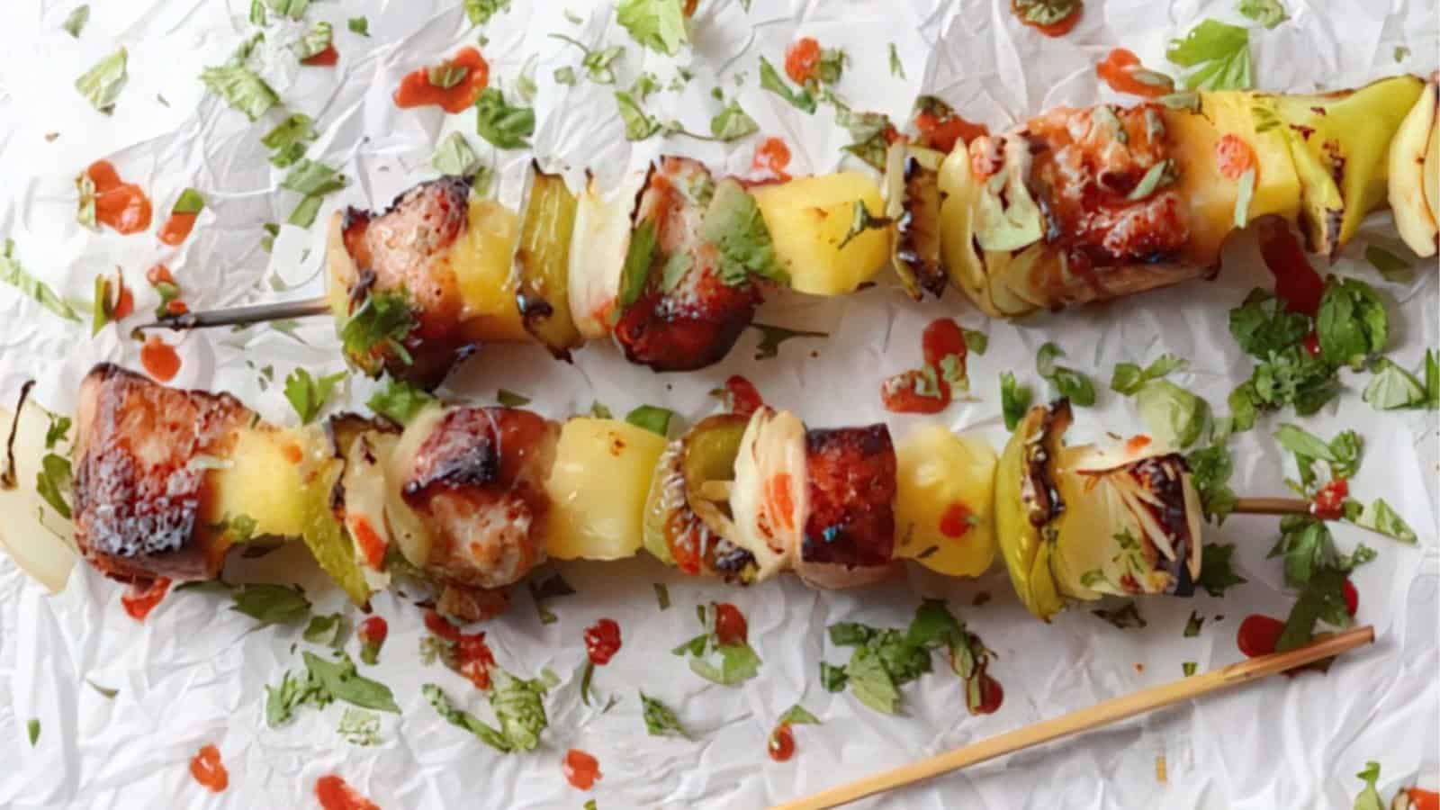 Grilled skewers with pieces of chicken, pineapple, onions, and green peppers, garnished with cilantro and a drizzle of hot sauce, placed on crumpled parchment paper.