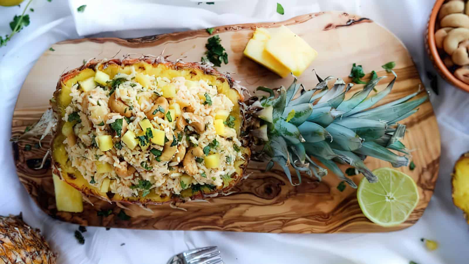 A halved pineapple is filled with a vibrant rice dish on a wooden platter. The dish is garnished with cashews, herbs, and pineapple chunks. A lime half and whole cashews are nearby. Bright and tropical presentation on a white background.