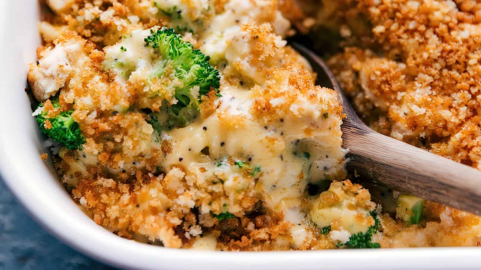 A close-up of a creamy broccoli casserole topped with golden breadcrumbs in a white dish. A wooden spoon is scooping a portion, revealing the rich, melted cheese sauce and vibrant green broccoli.
