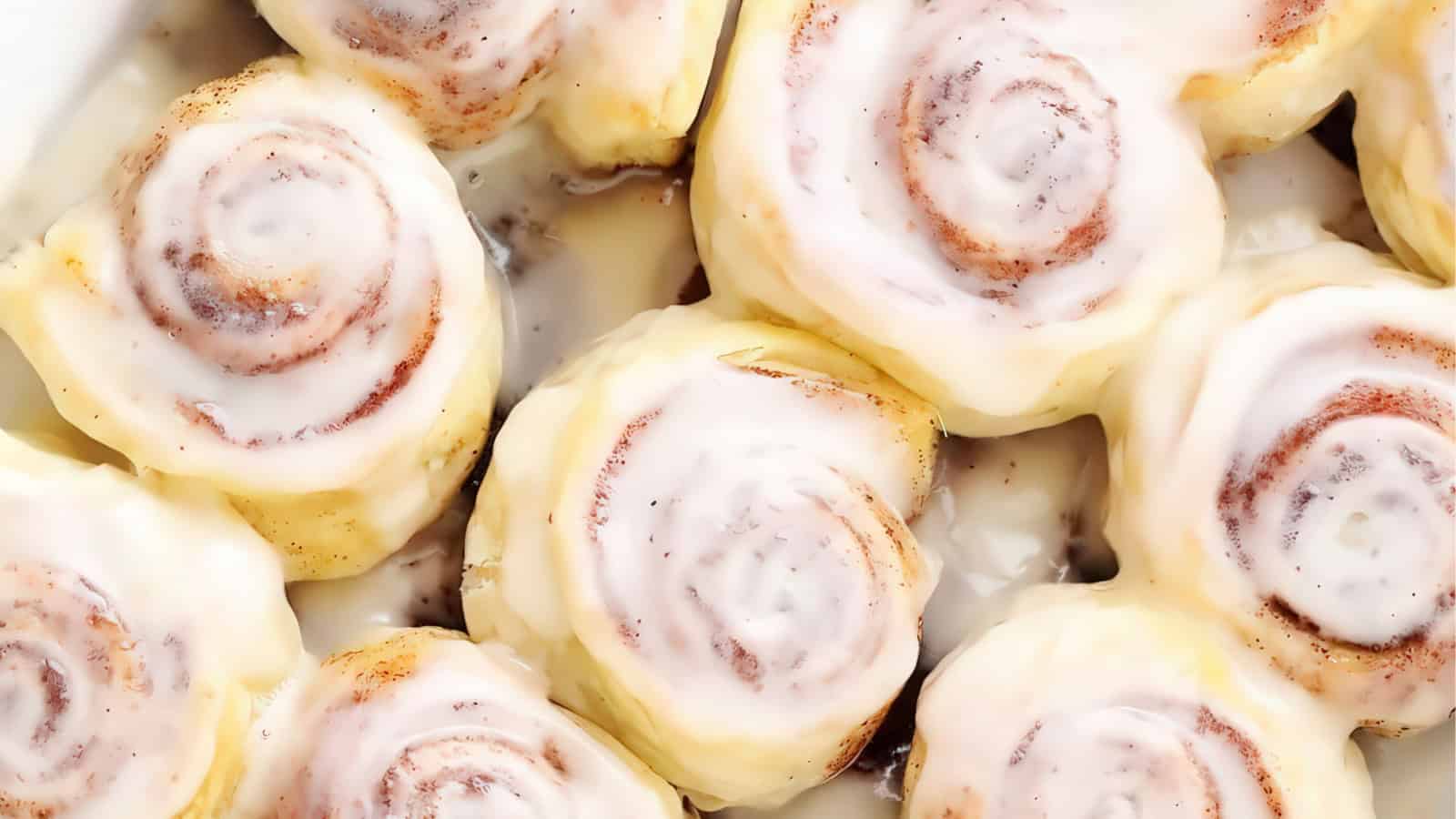 Close-up of freshly baked cinnamon rolls topped with creamy white icing, arranged closely together. The swirled layers of dough and cinnamon are visible under the glaze.