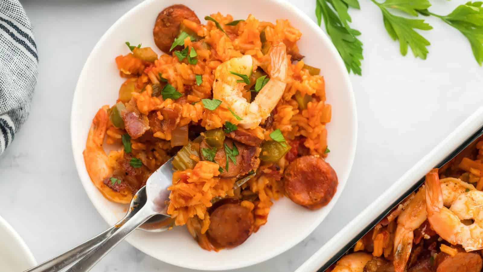 A plate of vibrant jambalaya with shrimp, sausage, and bell peppers, garnished with fresh parsley. A fork is placed on the side. Fresh parsley leaves are visible in the background.