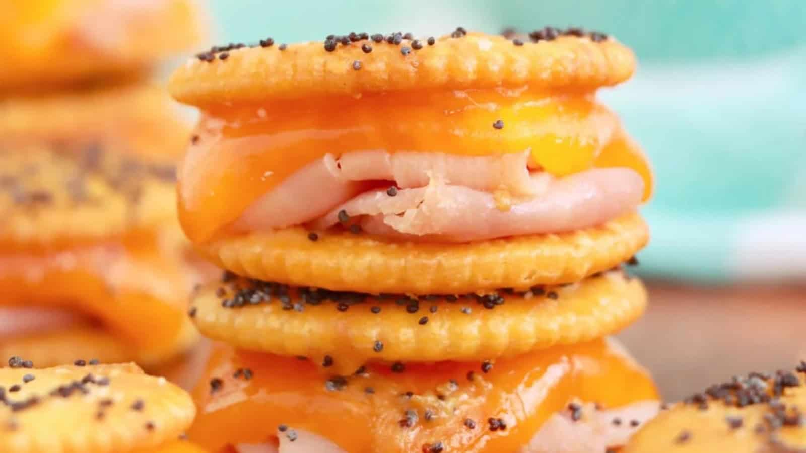 A stack of cheesy cracker sandwiches filled with turkey and melted cheddar cheese, topped with poppy seeds. The crackers are golden brown and slightly shiny from the cheese.