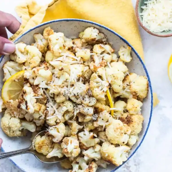A bowl of roasted cauliflower topped with cheese is accompanied by a lemon half and a small bowl of grated cheese on a yellow cloth. Hands are holding a spoon near the bowl.